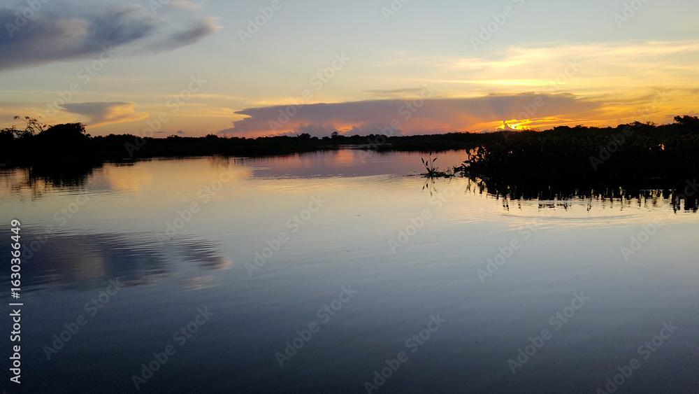 Fototapeta premium Stunning panoramic landscape of Cuiabá River in Pantanal wetlands at sunset with colorful clouds reflecting on water. The sun sets behind a silhouetted treeline, creating a peaceful natural scene.