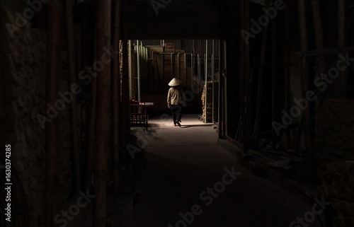  Evocative image captured inside a traditional incense village near Hanoi, Vietnam. A man wearing a conical hat walks through a dramatic beam of light inside a shadowy warehouse.