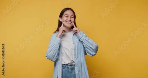 smiling young woman playfully points to her own cheeks, expressing joy and whimsy on vibrant yellow background