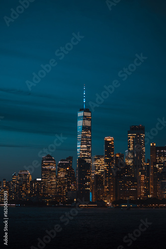 The illuminated Lower Manhattan skyline and One World Trade Center glittering at night, seen from the water.