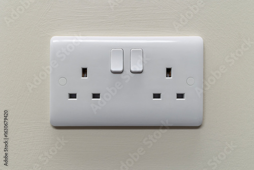 UK dual 3-pin 240V mains electrical socket with two individual switches. White plastic faceplate mounted on painted interior wall. Ideal for illustrating home or office electrical fittings