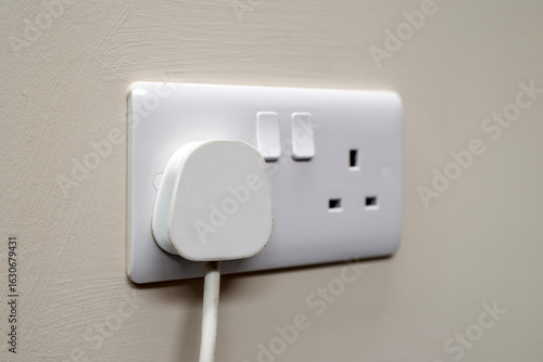 White UK 3-pin mains plug inserted into modern dual wall socket with switches. Mounted on a light painted wall. Suitable for illustrating household electricity, safety, and electrical infrastructure