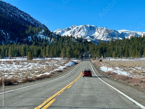 Straight highway stretching toward snow-covered mountains under clear blue sky, June Lake, California. Red SUV drives down center of empty road through winter landscape with scattered snow