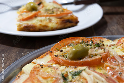 Part of an Argentinian Neapolitan pizza on a tray and a slice on a plate in the out-of-focus background
