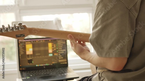 Musician composing music at home, working on laptop with DAW and playing the blue electric guitar. Creative music production process in cozy daylight home studio.