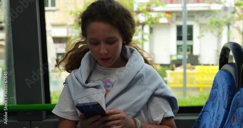 Young woman intently looks at phone sitting with back to window during bus trip. Teenager girl passenger concentrates attention on gadget in arms