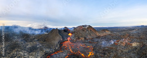 Aerial view of fiery lava flows through jagged, dark volcanic rock towards distant snowy peaks under a pale sky, Grindavik, GrindavÃ­kurbÃ¦r, Iceland.