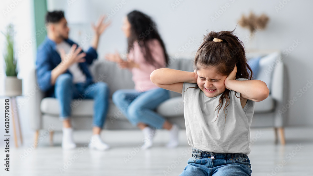 Obraz premium Family Conflicts. Little arabic girl covering ears with hands while her parents arguing on background, upset child doesn't want to hear quarrel, stressed kid sitting on floor alone, selective focus
