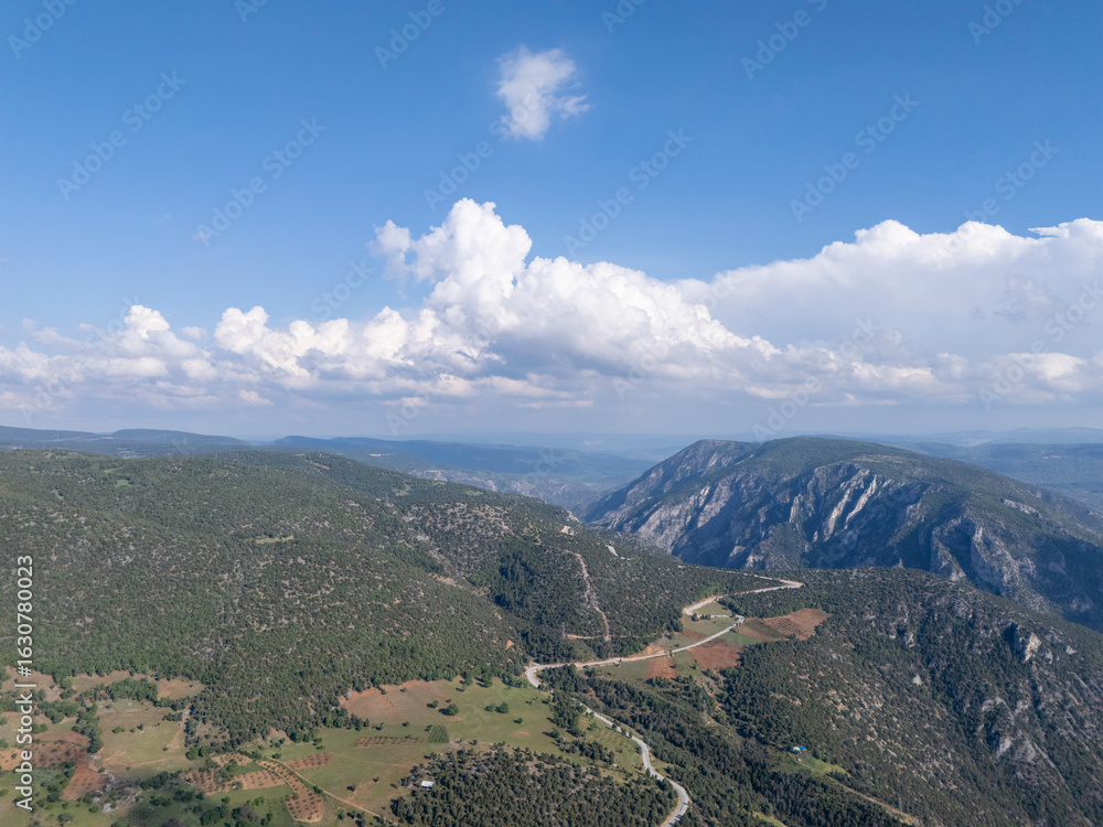 Fototapeta premium Scenic landscape of Gelemiç, Bursa, featuring lush green hills, winding roads, rocky mountains, and a vibrant blue sky with fluffy clouds, capturing the serene beauty of nature. 