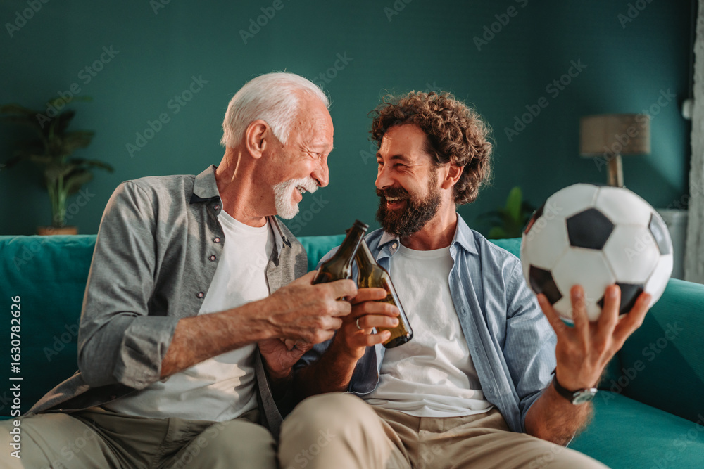 Fototapeta premium Father and son celebrating with beer bottles on sofa at home