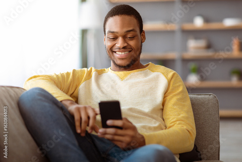 фотографія Cheerful black guy reclining on couch at home, using mobile hone, chatting with friends or playing online games, copy space