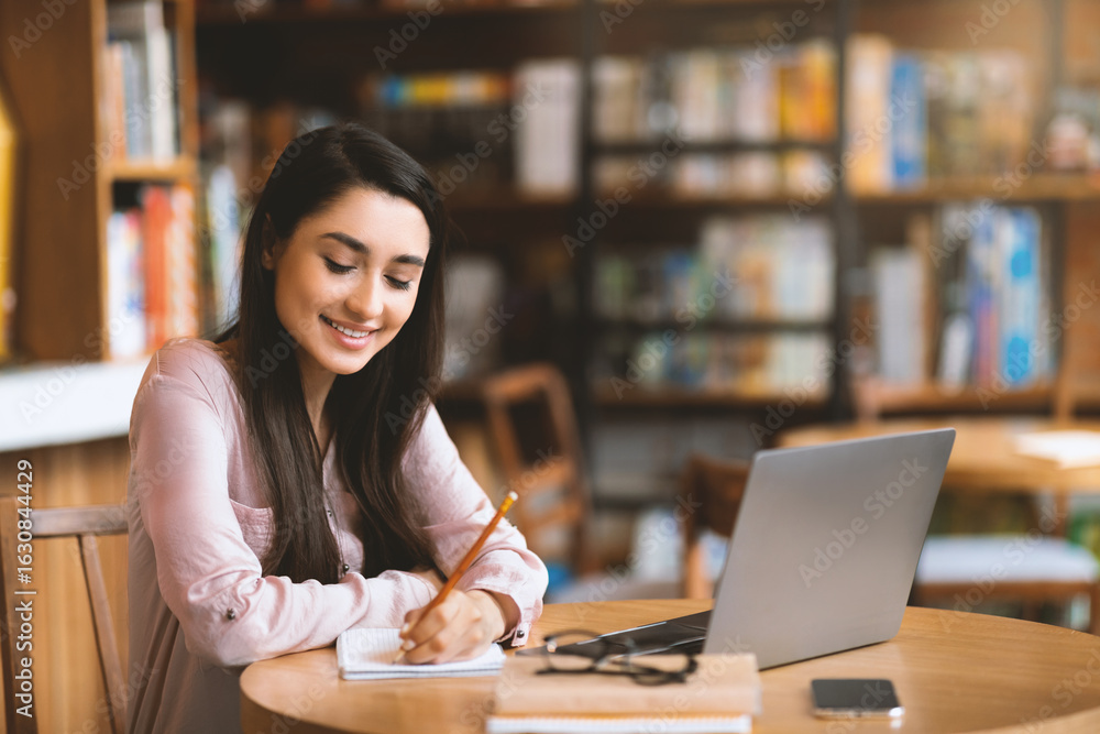 Online education. Motivated latin woman studying with laptop at cafe and taking notes while watching webinar or preparing for exam in cafeteria, empty space Online education. Motivated latin woman studying with laptop at cafe and taking notes while watching webinar or preparing for exam in cafeteria, empty space