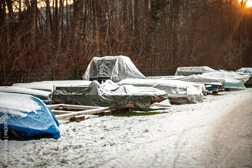 Obraz na plátne Row of small boats covered with tarps and dusted with snow in Austria, stored on land for the cold season beside a forest path