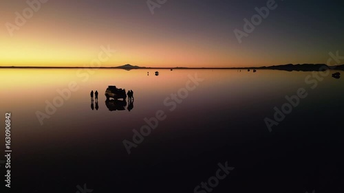 Drone footage circling 4x4 vehicle and two people on chairs at salt flats during sunset. Perfect mirror reflection on water surface. Mountains silhouetted on horizon at dusk.