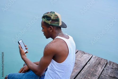 Afro Caribbean Man Sitting By The Dock  On A Rustic Deck With His Mobile Phone.