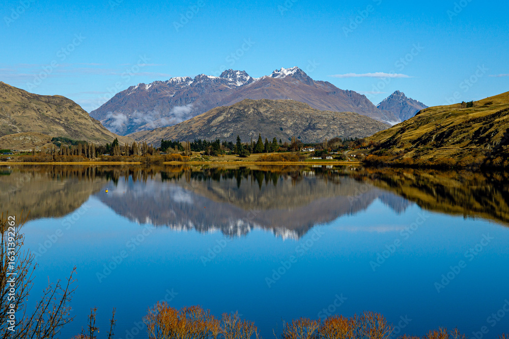 Obraz premium Snow-dusted mountains reflect perfectly in a calm lake under a clear blue sky near Queenstown, New Zealand, with trees and fields lining the peaceful shoreline.