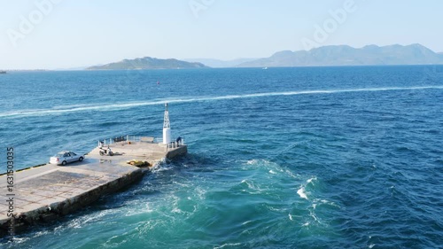 Wallpaper Mural A top-down view of a pier stretching into turquoise waters on Aegina Island, Greece. A white lighthouse stands at the edge, with a man nearby. A car and scooter are parked. Mountains in distance. Torontodigital.ca
