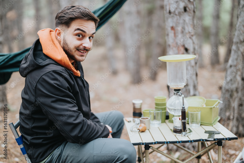 Fototapeta premium Outdoor camping scene with a man in a forest, sitting near a table set with coffee preparation utensils. The woodland background enhances the atmosphere of relaxation and adventure.
