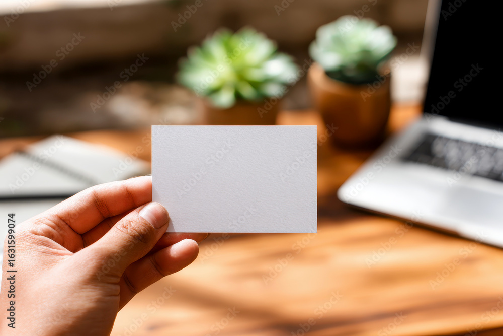 Obraz premium Hand holding blank business card mockup in cozy workspace with notebook. White card template against marble table background. Perfect for branding presentation.