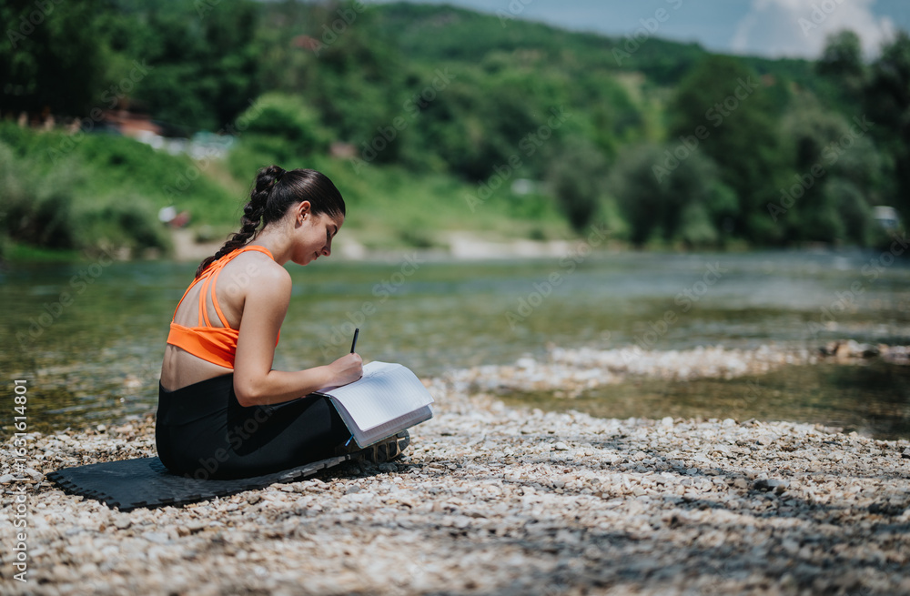 Fototapeta premium A young woman sits by the river on a sunny day, writing in her journal. The tranquil surroundings and peaceful environment create a perfect atmosphere for relaxation and reflection.