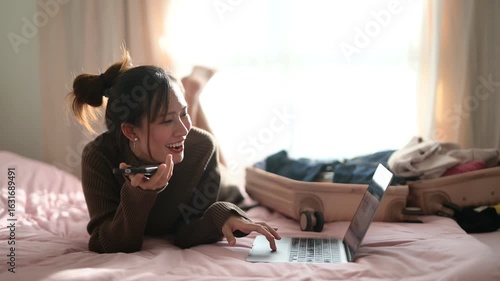 A woman is lying on a bed, smiling while talking on her phone and using a laptop, with luggage and clothes visible nearby.