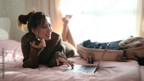 A woman is lying on a bed, smiling while talking on her phone and using a laptop, with luggage and clothes visible nearby.