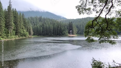 Misty Synevyr Lake in the Carpathian Mountains on a rainy day. Tranquil water, forest landscape, and green hills in the background.