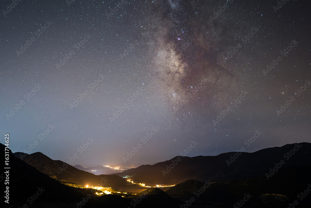 Fototapeta premium Milky Way galaxy above mountains and glowing village lights at night