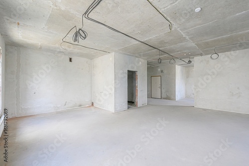 An unfinished, empty room with concrete walls and ceiling, visible wiring, and doorways. The space awaits finishing touches and renovation
