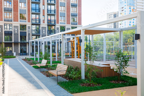 A modern residential complex with a landscaped area: playground, benches, pergola, green spaces, neat paths, cozy courtyard, brick facades, glass windows.