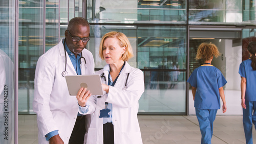 Male And Female Doctors Having Informal Meeting In Modern Hospital Looking At Digital Tablet