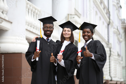 Portrait of a group of smiling happy multiracial international graduates students hugging and having fun in a university graduate gown and holding diploma outdoor. Education and graduation concept.