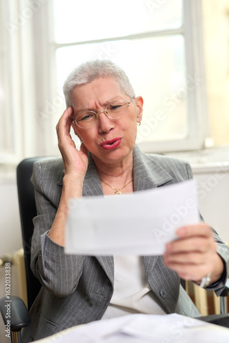 An older woman with short gray hair seated in an office chair looks stressed while holding documents. She appears to be feeling overwhelmed by the content and is deep in thought