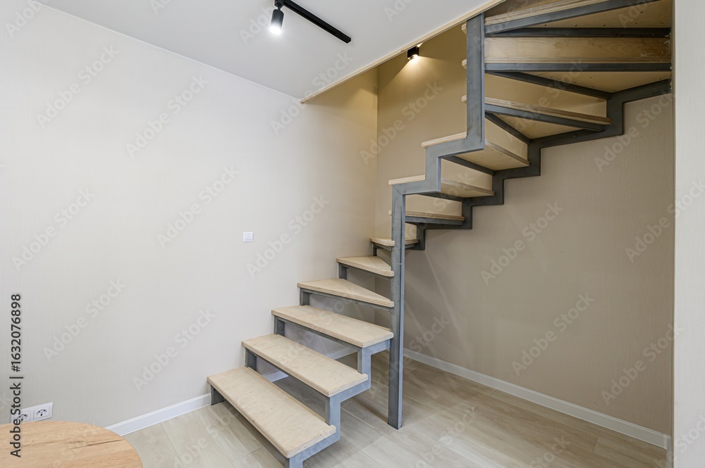 Fototapeta premium modern staircase with wooden steps and a metal frame stands in a room with beige walls and light wood floors. Black track lighting is on the ceiling