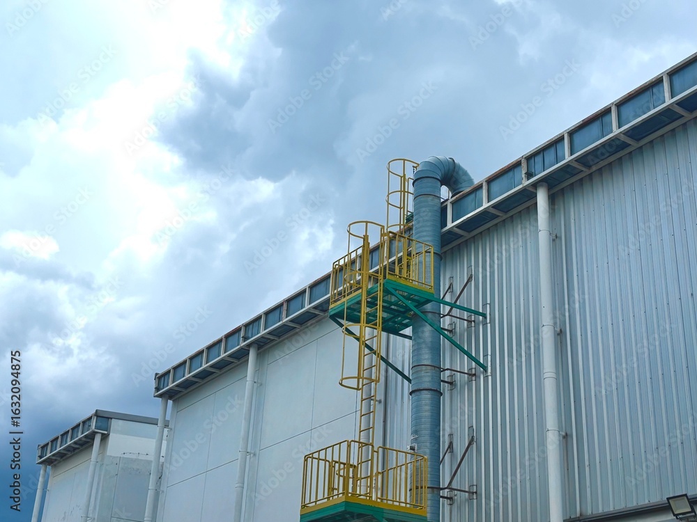 Fototapeta premium A large industrial building with a ventilation system and a maintenance ladder against a dramatic, cloudy sky. This image represents modern factory infrastructure and environmental control.