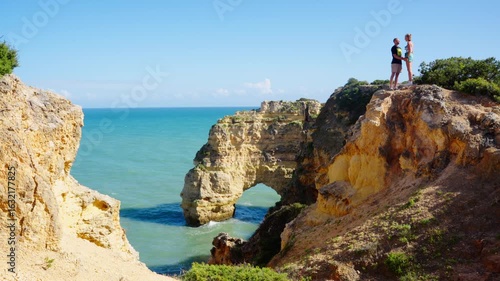 A Couple Over A Rock Cliff Arch Near Marinha Beach In Algarve, Portugal. Wide Shot