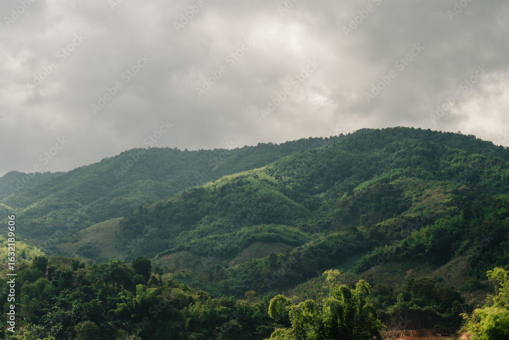 Obraz premium Rolling green hills and mountains under a cloudy, overcast sky. Lush vegetation covers the slopes.