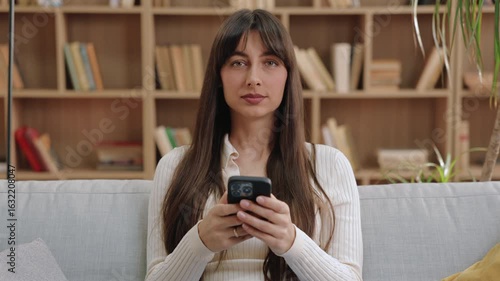 Serious focused Caucasian woman sitting still on sofa and holding smartphone with both hands. Staring ahead or listening attentively during video call in quiet room filled with bookshelves.