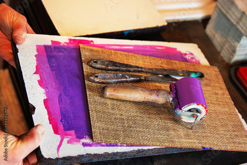 Hands of artist holding a textured printing plate with a purple inked brayer and palette knives arranged on top. Printmaking or monotype printing tools
