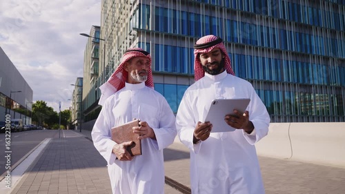 Two emirati businessmen wearing traditional saudi arabia clothing are posing together in front of modern glass buildings, creating a striking juxtaposition of tradition and modernity