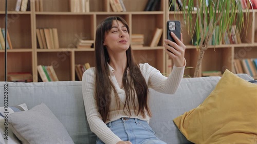 Smiling beautiful Caucasian woman sitting on couch holding smartphone and waving hand. Having video call or chatting with friend in bright cozy room with pillows and wooden bookshelf behind.