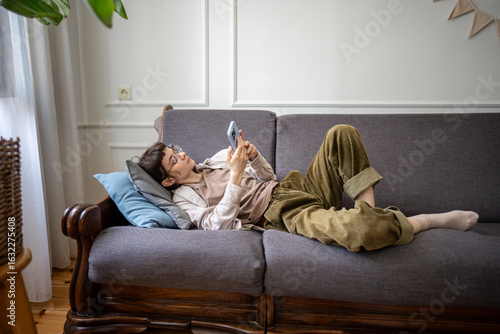 Fotografie Procrastinating young woman lounging on sofa browsing chat apps on smartphone, laziness and emotional detachment