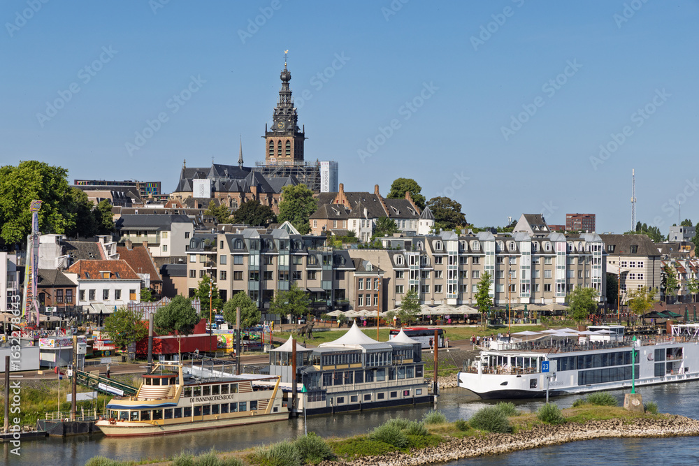 Fototapeta premium The historic city of Nijmegen, Netherlands, seen from the riverside with the prominent St. Stephen's Church and boats docked along the quay. Nijmegen, Netherlands. 20 June 2025.