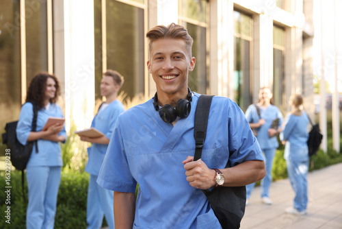 Obraz na plátne Happy medical student in uniform with backpack and headphones near building outd