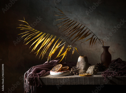 Still life with a dry palm branch in a clay vase and dried oranges on a dark background