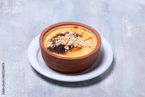 High angle studio photo of baked milk pudding with crcushed hazelnut topping  in earthenware pot, a traditonal Turkish dessert called fırında sütlaç on white wooden table