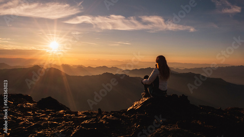 A woman sits on a rocky peak, silhouetted against a sunrise over distant mountains. The sun bursts through the clouds, casting rays across the landscape. Mantova refuge al Vioz, Peio, Trentino.