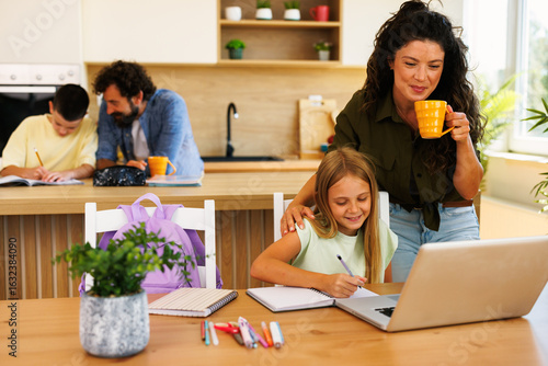 Family enjoying time together doing homework and homeschooling with computer while mother drinks coffee