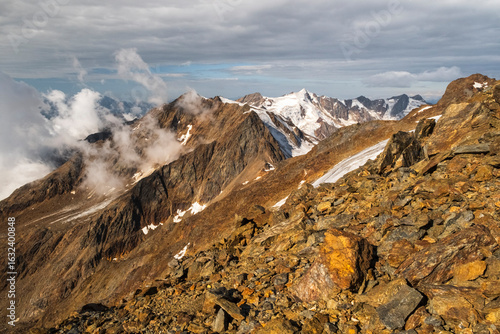 Aerial view of rocky mountain peaks, some covered in snow and partially obscured by clouds, with glaciers visible in the distance. View from Mantova refuge al Vioz, Peio, Trentino. 