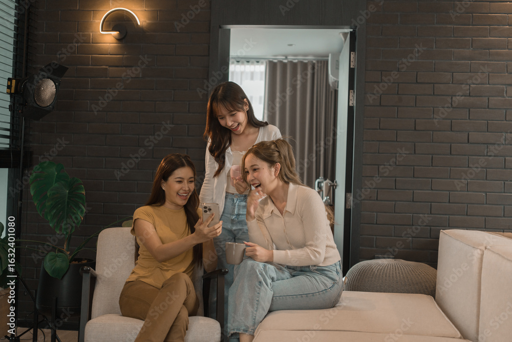 Obraz premium Three young Asian women shopping online at home using a laptop and credit card. They are smiling and enjoying time together in a relaxed and modern living room.
